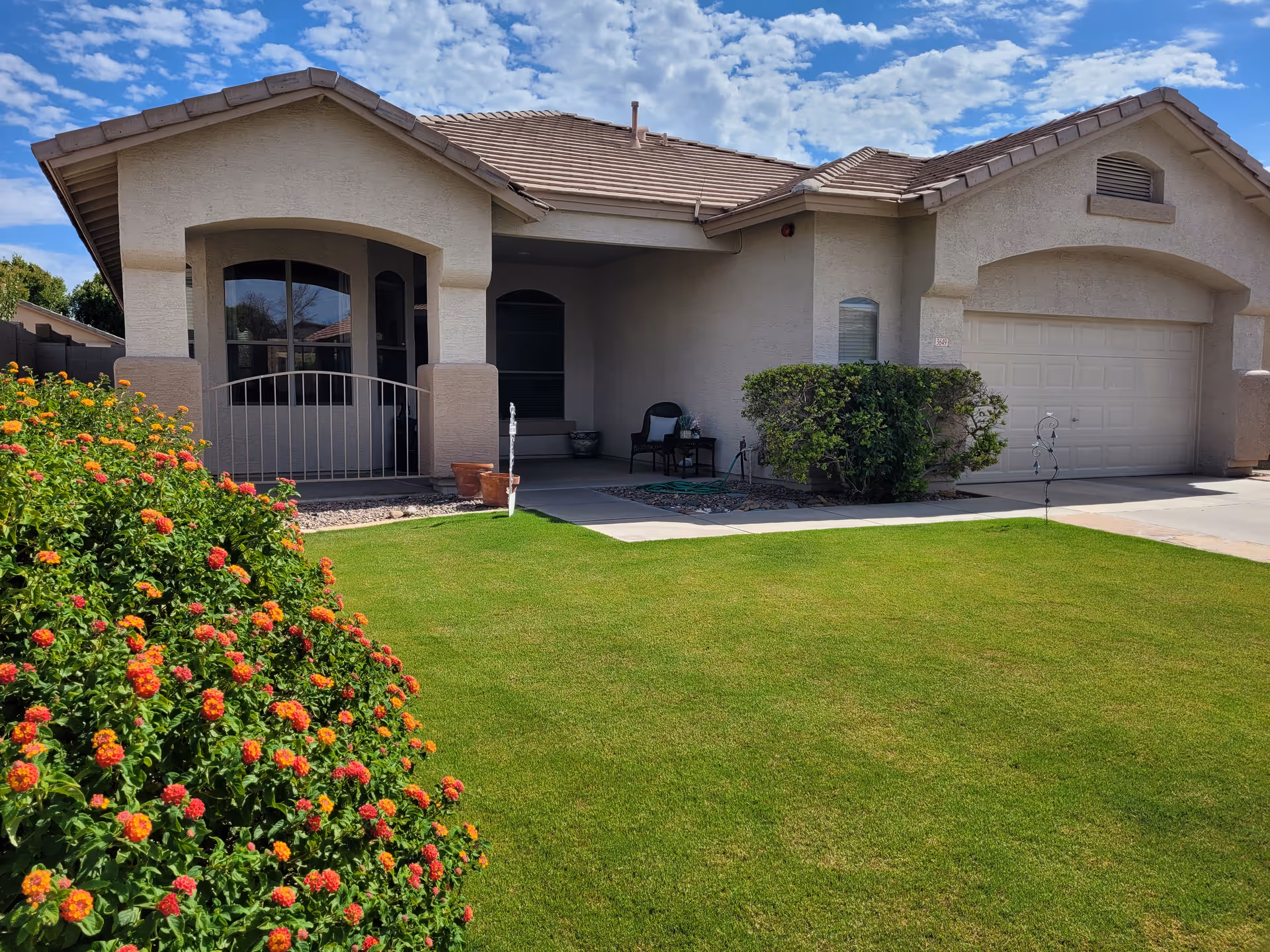 Front exterior view of a single-story house with a tiled roof, beige stucco walls, a two-car garage, a covered porch with seating, a well-maintained green lawn, and flowering bushes in the foreground under a partly cloudy blue sky.