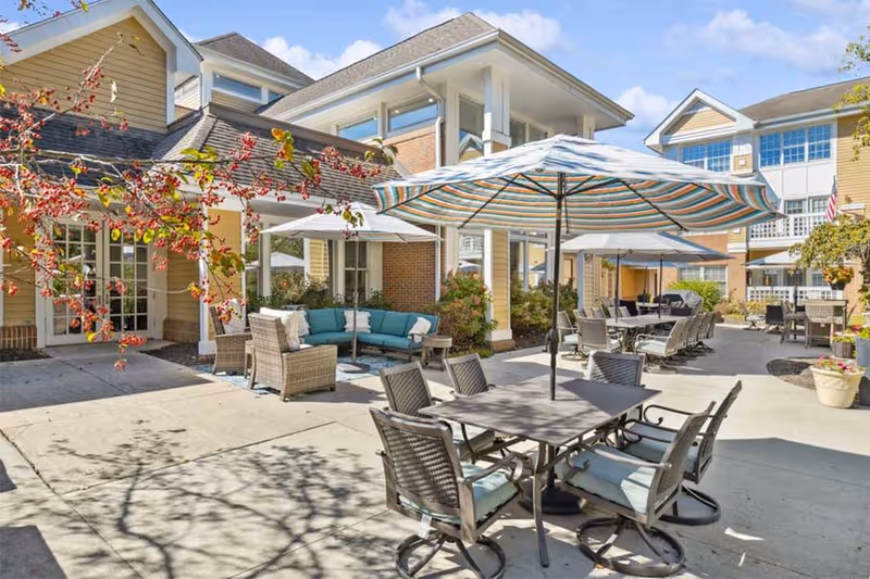 Outdoor patio area at a senior living facility with multiple tables and chairs, some shaded by large striped umbrellas. There is a seating area with a blue cushioned sectional sofa and wicker chairs near the building. The building has yellow siding and large windows, and the sky is clear with a few clouds.