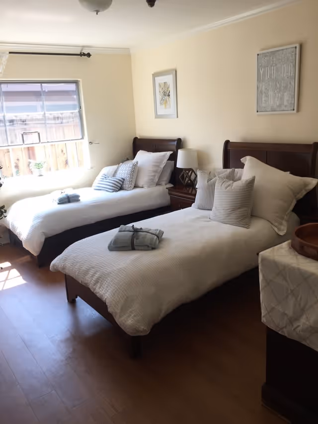A bright bedroom with two single beds made up with white bedding and multiple pillows. There is a wooden nightstand with a lamp between the beds. The room has light-colored walls with two framed pictures hanging above the beds. A window on the left side lets in natural light, and the floor is wooden.