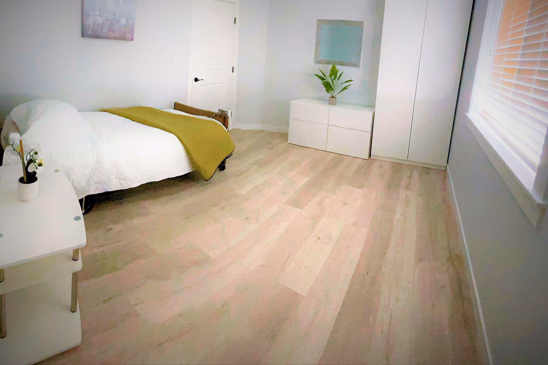 A bright, minimalist bedroom with light wood flooring, a single bed with white bedding and a green blanket, a small white table with a potted plant, a white dresser with a green plant on top, a white wardrobe, a window with blinds, and a closed white door.