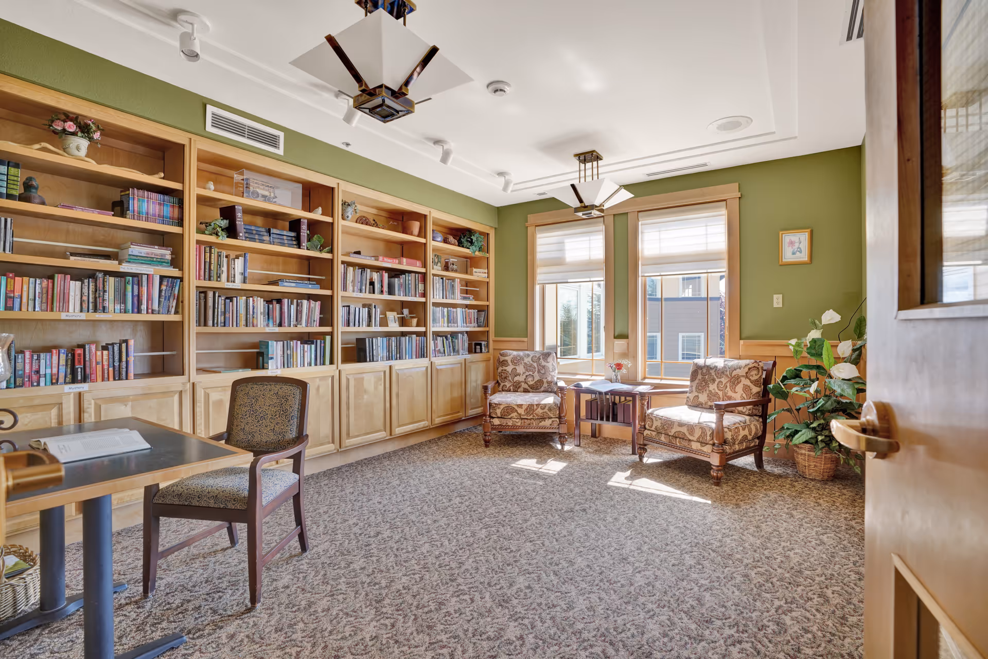 A cozy reading room with large wooden bookshelves filled with books along one wall, two upholstered armchairs with patterned fabric near two windows letting in natural light, a small table between the chairs, a single chair at a small table on the left, green walls with wood trim, and carpeted floor.