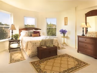 A bright and cozy bedroom with a large bed adorned with floral bedding and multiple pillows. The room features three windows letting in natural light, a wooden nightstand with a lamp and plant, a small round table with a vase of purple flowers, a wooden dresser with a mirror and lamp, and two patterned area rugs on the carpeted floor.