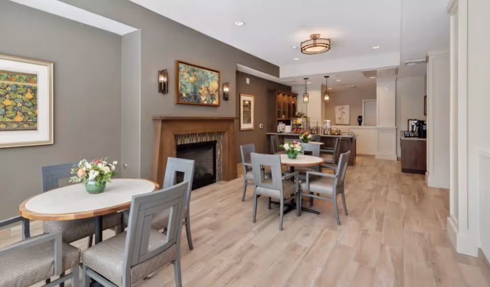 Bright communal dining area with round tables and chairs, a fireplace, and a kitchen/bar area in the background.
