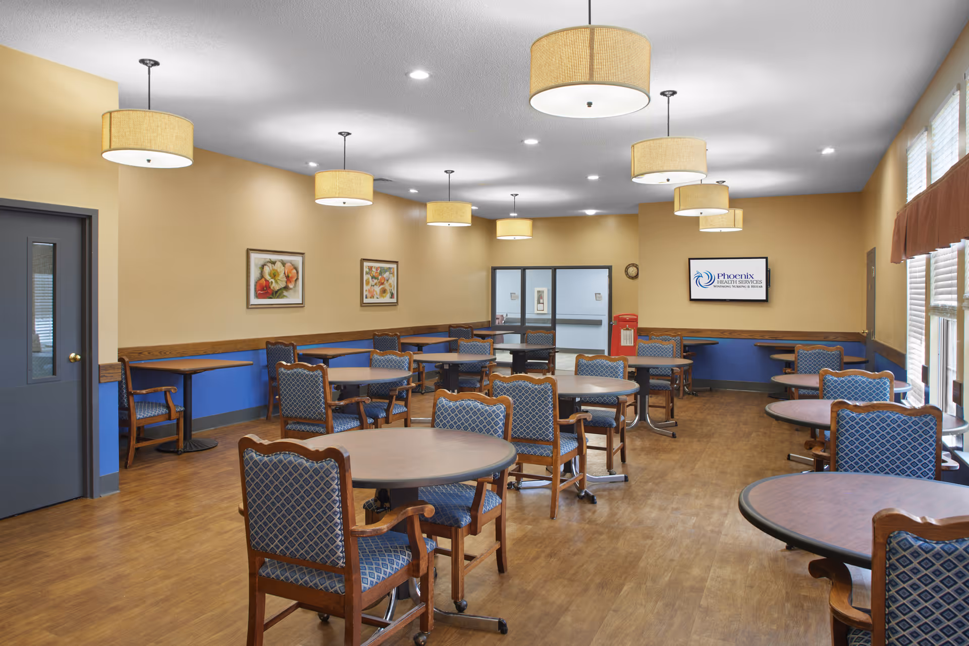 Spacious dining room with round tables, patterned chairs, pendant lights, and large windows in a senior living facility.