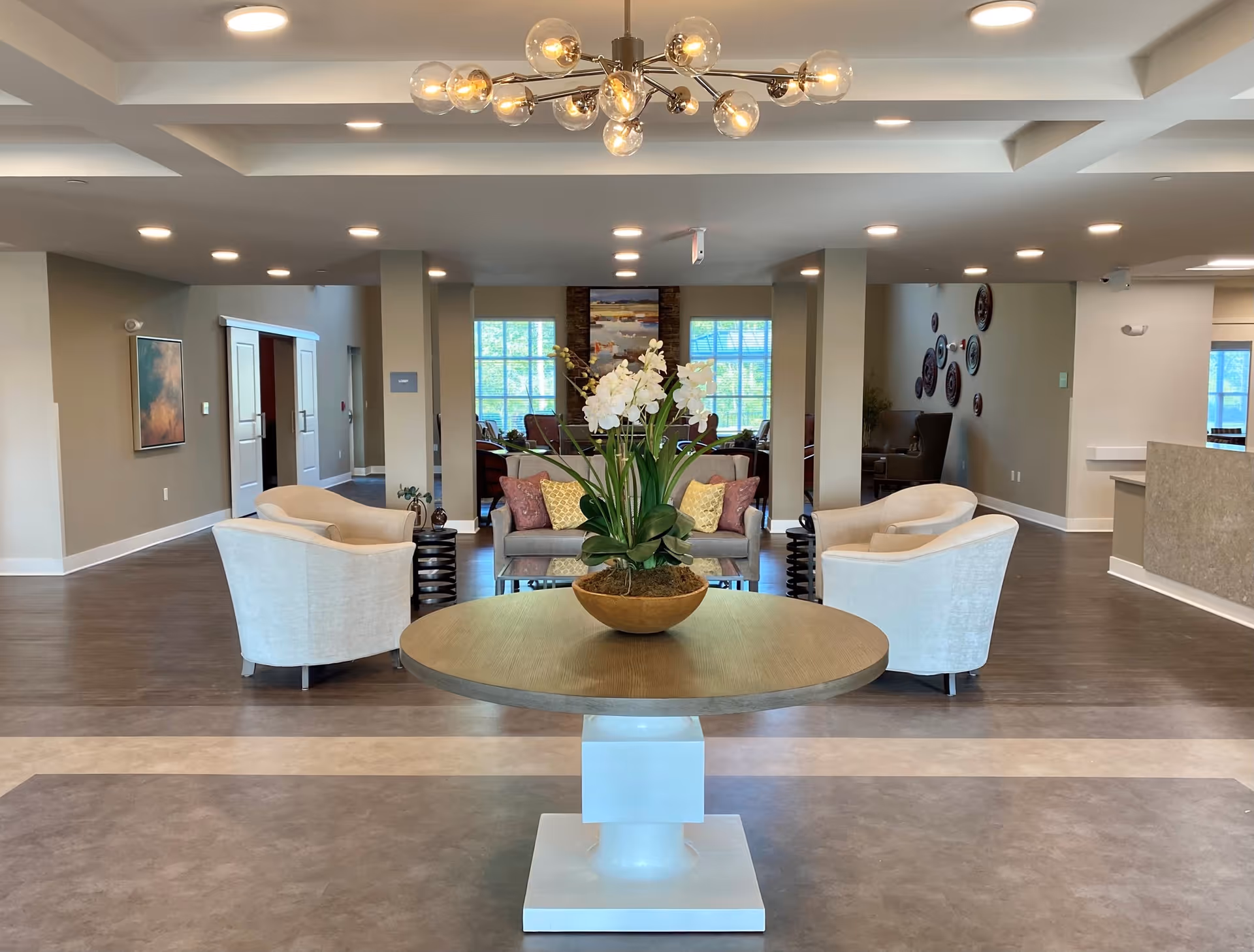 A spacious and well-lit common area in a senior living facility featuring a round wooden table with a large flower arrangement in the center. Surrounding the table are four white upholstered armchairs arranged in pairs, with a sofa and additional seating visible in the background. The room has a modern chandelier, recessed ceiling lights, and large windows allowing natural light to enter.