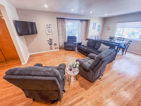 A spacious living room area with wooden flooring featuring three gray recliner chairs arranged around a small round table with a flower vase and magazines. A large gray sofa with pillows is placed near a window with curtains. A flat-screen TV is mounted on the wall to the left. In the background, there is a dining area with a table and chairs near a window with blinds.