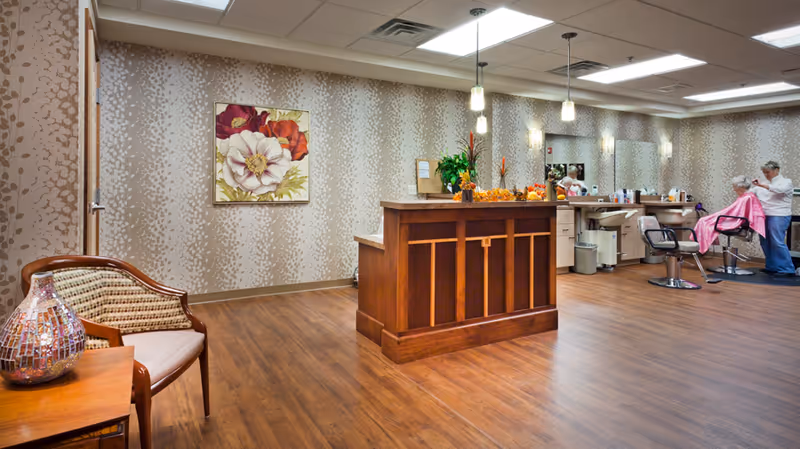 Interior of a senior living facility's hair salon area with a wooden reception desk in the center, a floral painting on the wall, a chair with a decorative vase on a small table to the left, and a hairstylist cutting an elderly woman's hair on the right side. The room has wood flooring, patterned wallpaper, and bright overhead lighting.