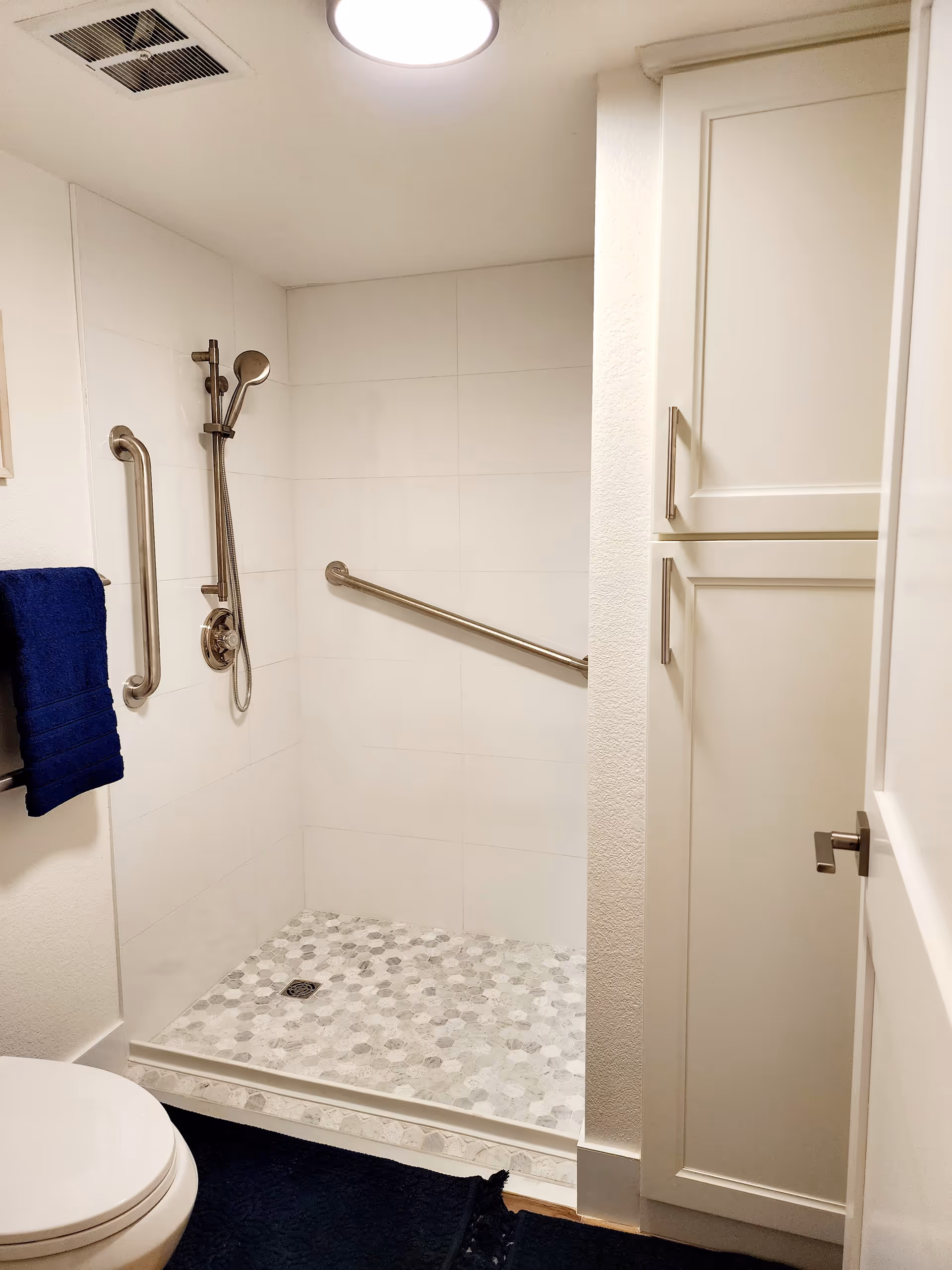 A bathroom with a walk-in shower featuring white tiled walls and a hexagonal patterned floor. The shower has a handheld showerhead and two stainless steel grab bars for support. A white toilet is partially visible on the left, and a white cabinet with silver handles is on the right. A navy blue towel hangs on a rack near the shower, and a navy blue rug is on the floor.