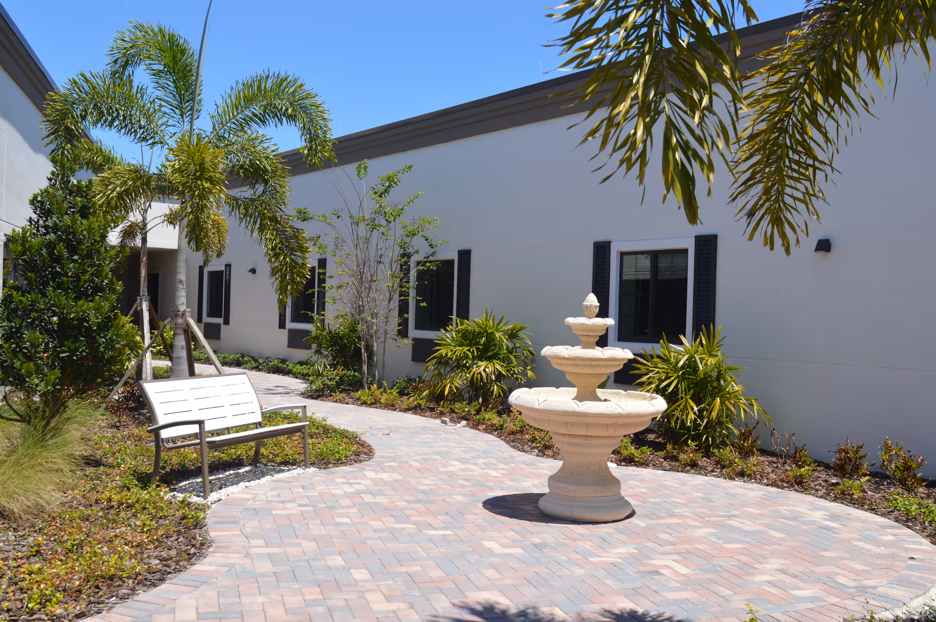 Sunlit courtyard with a three-tier stone fountain, a bench, palm trees, and a white building facade.