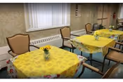 Small dining room with tables covered in yellow tablecloths and chairs, each table decorated with a vase of sunflowers.