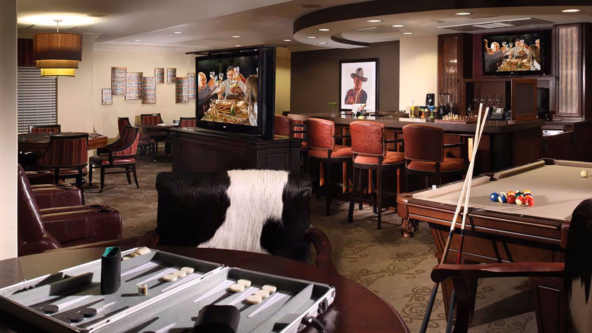 A cozy game and lounge room featuring a backgammon board on a table in the foreground, a pool table with cues and balls, a bar area with red cushioned stools, a TV mounted above the bar showing people toasting, and a dining area with tables and chairs in the background. The room has warm lighting and decorative wall art.