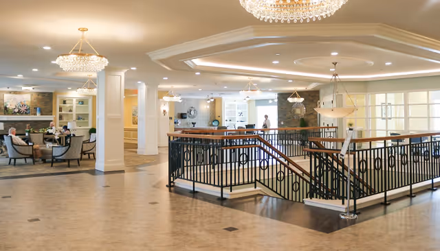 Spacious and well-lit interior common area of a senior living facility featuring a central staircase with black railings, elegant chandeliers, and comfortable seating areas where elderly residents are sitting and conversing. The space has light-colored walls, tiled flooring, and decorative elements including artwork and plants.