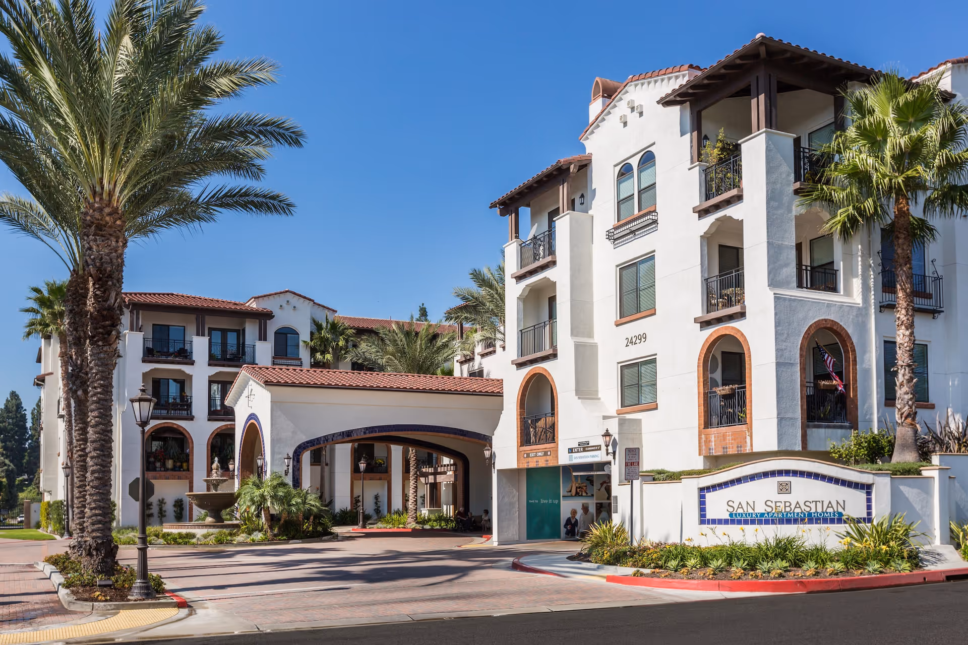 Exterior view of the San Sebastian luxury apartment homes building with white walls, red-tiled roof, balconies, palm trees, and a covered entrance driveway under a clear blue sky.
