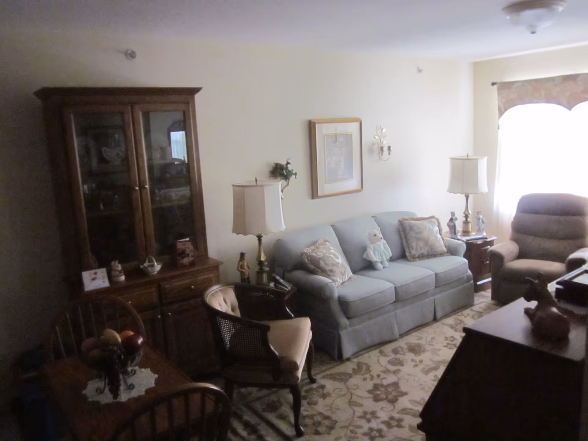Cozy living room with a blue upholstered sofa (with a teddy bear), armchair, wooden china cabinet, side tables and lamps.