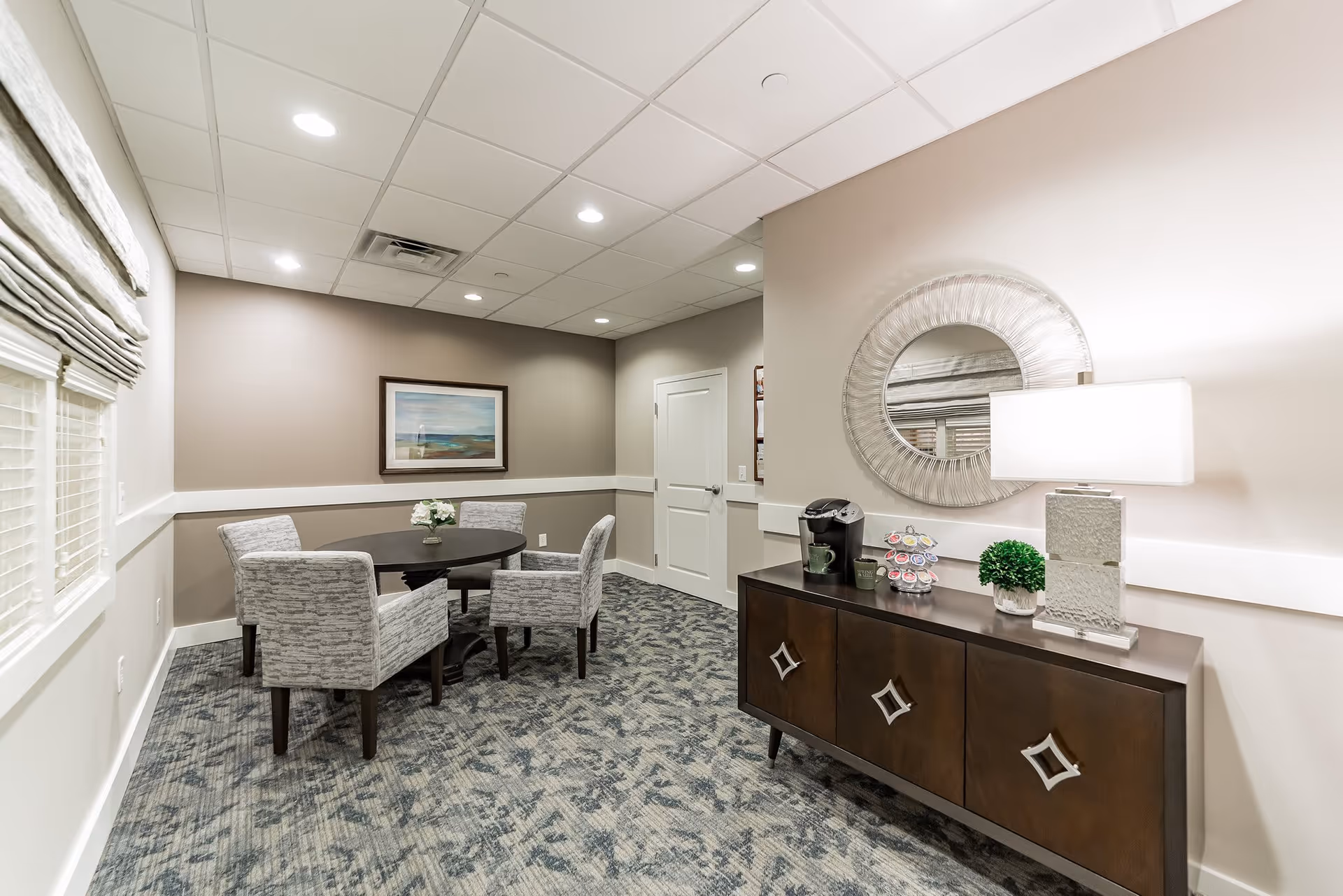 A small, well-lit room with a round table surrounded by four upholstered chairs. On the right side, there is a dark wooden sideboard with a table lamp, a coffee maker, a small plant, and a coffee pod holder. A round decorative mirror hangs above the sideboard. The walls are painted in neutral tones, and there is a framed landscape painting on the far wall. The floor is carpeted with a patterned design, and a window with blinds is on the left wall.