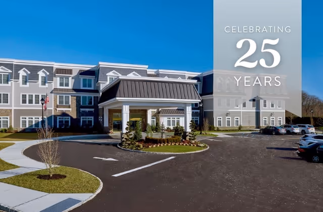 Front entrance of a three-story assisted living building with a covered drop-off, surrounding driveway and parked cars, and a banner reading 'Celebrating 25 Years'.