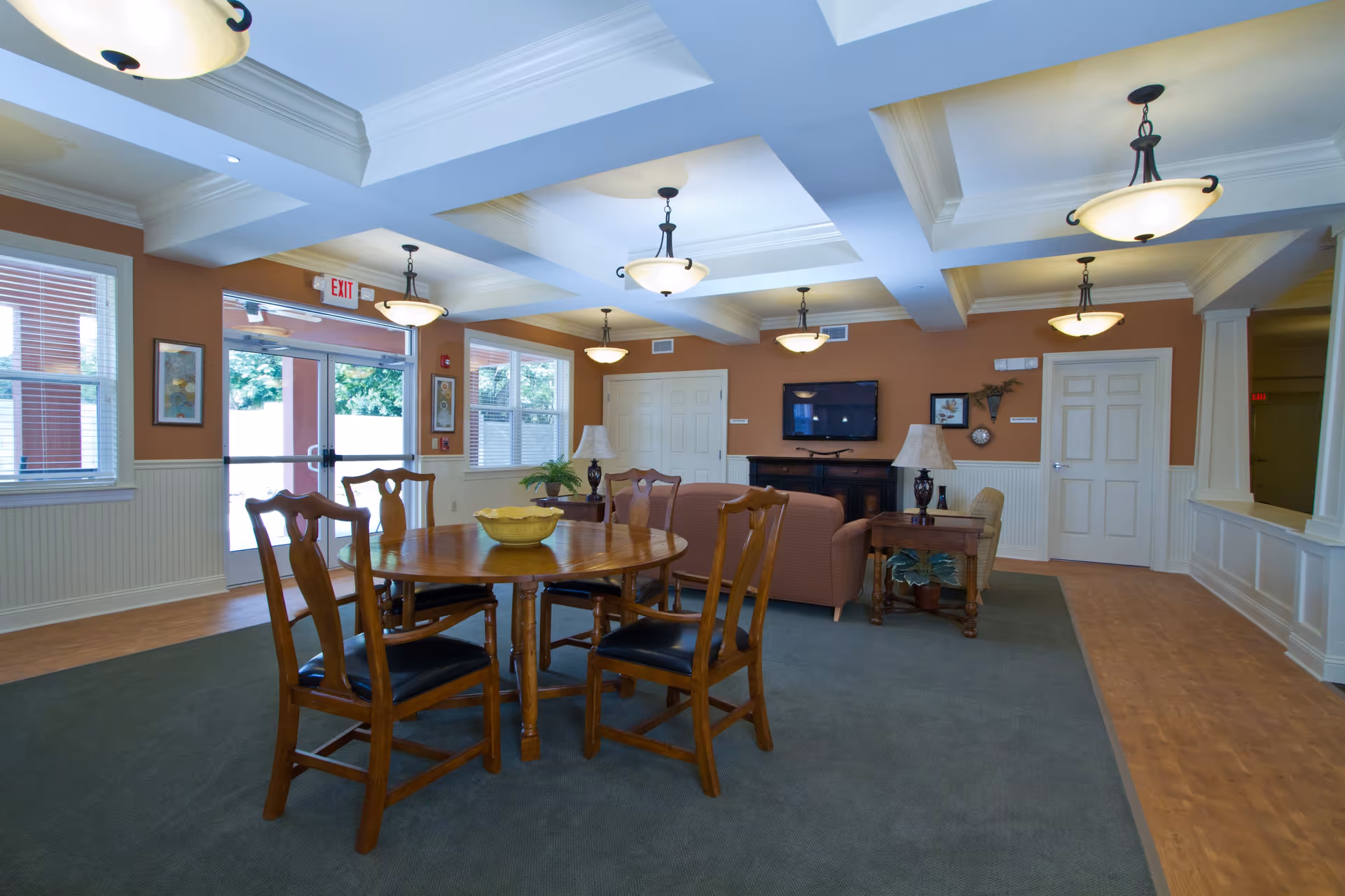 A spacious and well-lit common area with a round wooden table and four chairs in the foreground. The room features a coffered ceiling with multiple hanging light fixtures, a seating area with a sofa and armchair, two table lamps, a flat-screen TV mounted on the wall, and large windows letting in natural light. The walls are painted a warm beige color, and the floor is a combination of carpet and wood.
