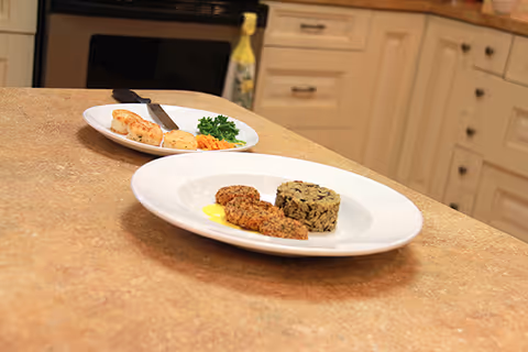Two white plates with food on a kitchen countertop. One plate has a serving of rice and three breaded pieces with a yellow sauce, and the other plate has biscuits, shredded cheese, and a sprig of parsley. Kitchen cabinets and an oven are visible in the background.