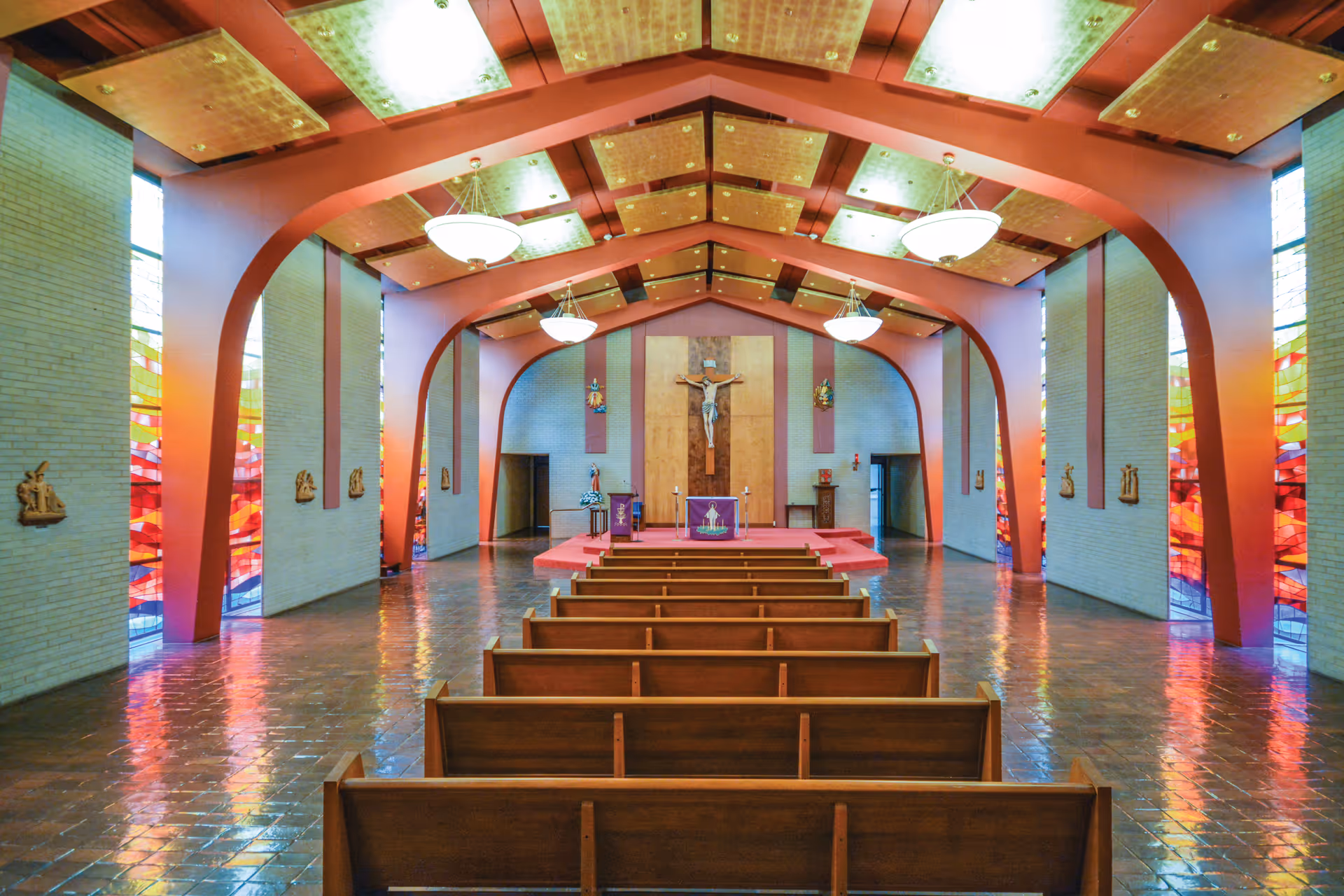 Interior view of a chapel with rows of wooden pews facing an altar, crucifix, and colorful stained glass windows.