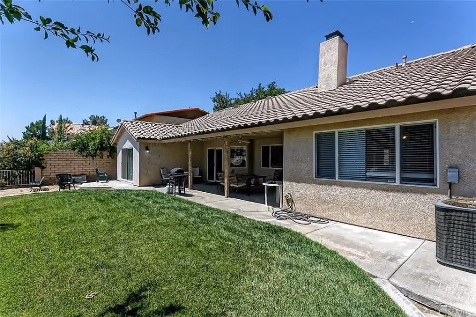 Backyard view of a single-story house with a tiled roof, beige stucco walls, and a covered patio area with outdoor furniture and a barbecue grill. The yard has a well-maintained green lawn and a concrete walkway along the house. Trees and a brick wall are visible in the background under a clear blue sky.