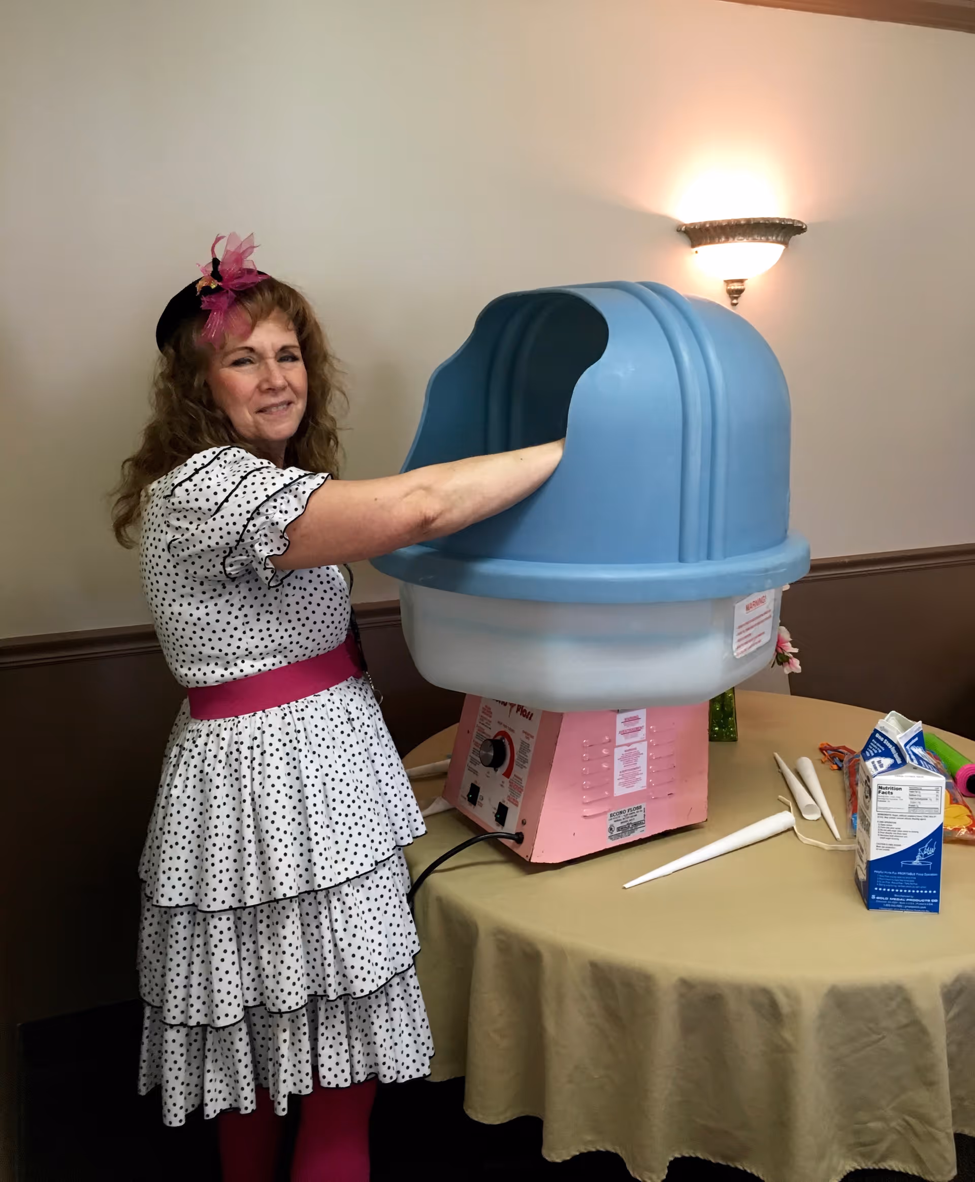 A woman in a white dress with black polka dots and a pink belt is standing next to a cotton candy machine with a blue cover, making cotton candy. The machine is on a round table covered with a beige tablecloth, and there are some supplies and a carton on the table. The background shows a wall with a sconce light.