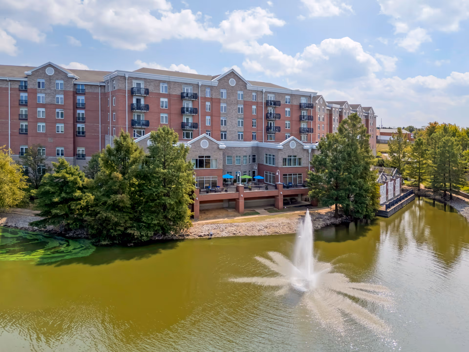 A large multi-story senior living facility building with balconies overlooking a pond with a water fountain. The building is surrounded by trees and has a partly cloudy sky above.