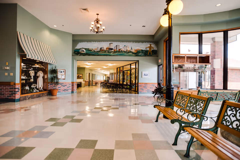 A spacious interior hallway with patterned tile flooring and green walls with brick accents. On the right side, there are decorative wooden benches with green metal armrests and a tall black lamp post. Large windows allow natural light to fill the space. At the end of the hallway, there is a room with tables and chairs visible through glass walls. Above the entrance to this room is a mural depicting a city skyline and a yellow figure.