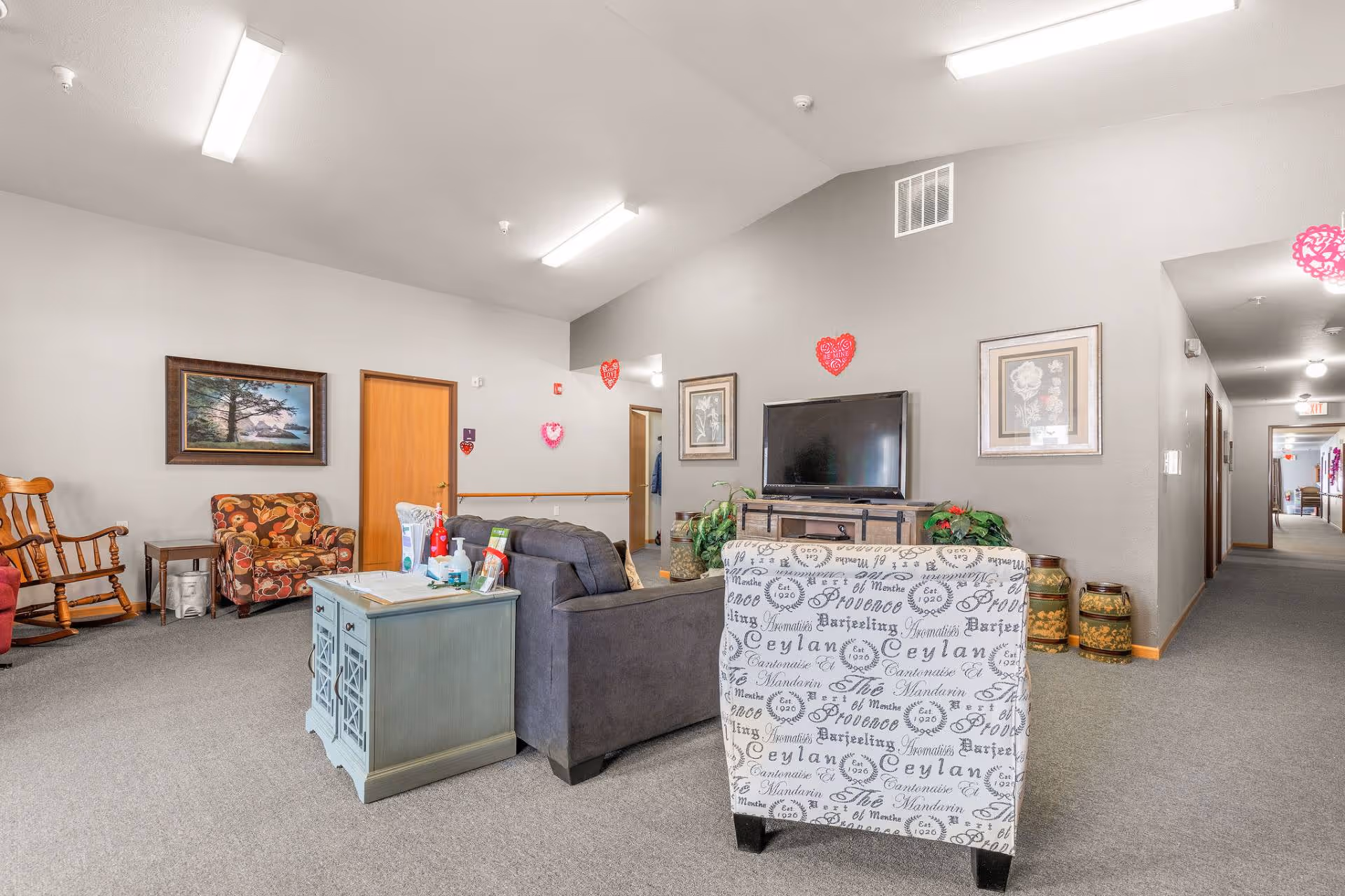 A cozy living room area in a senior living facility with a gray sofa, a patterned armchair, a wooden rocking chair, and a floral armchair. There is a TV on a wooden stand against the wall, framed artwork, and heart-shaped decorations on the walls. A hallway extends to the right with doors along it.