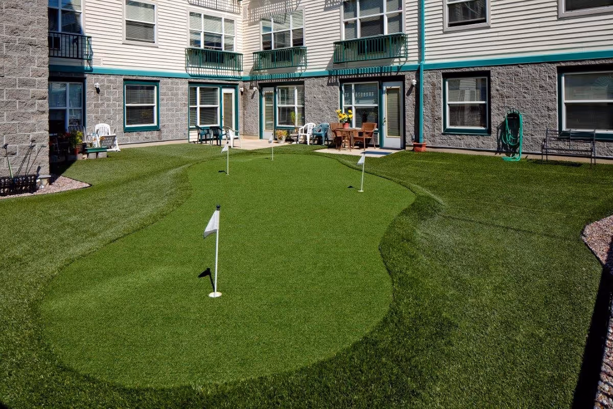 Outdoor courtyard area of Hazelwood Community Apartments featuring a small artificial putting green with four holes and flags, surrounded by green artificial turf. The courtyard is enclosed by a multi-story building with windows and doors, some with small balconies. There are chairs and a table with flowers near the doors.