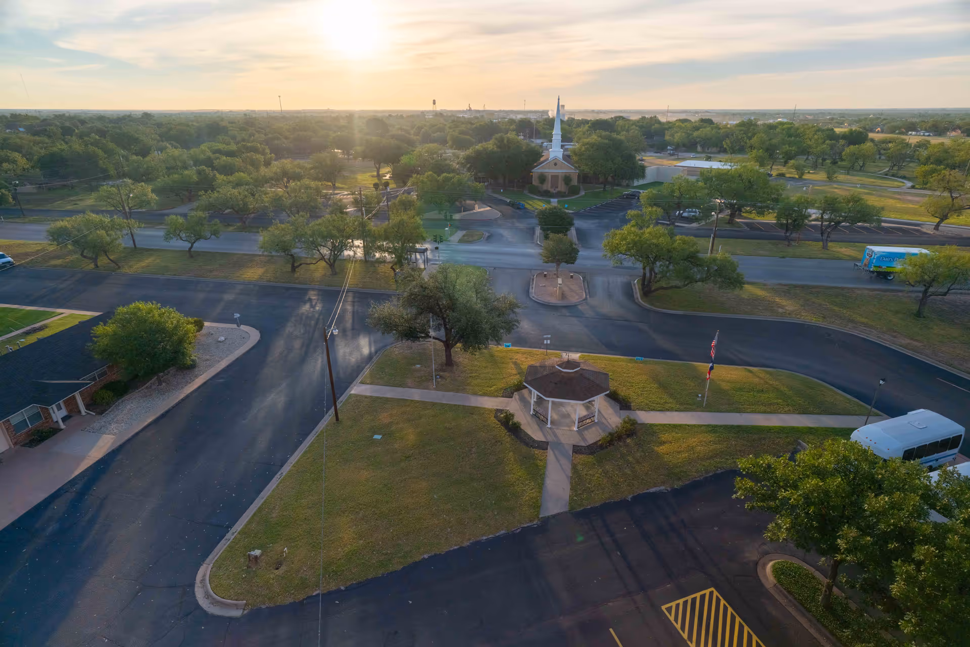 Aerial view of a retirement community area at sunset featuring a small gazebo on a grassy patch, a church with a tall white steeple in the background, several trees, paved roads, parking areas, and a few vehicles including a white van and a blue truck.