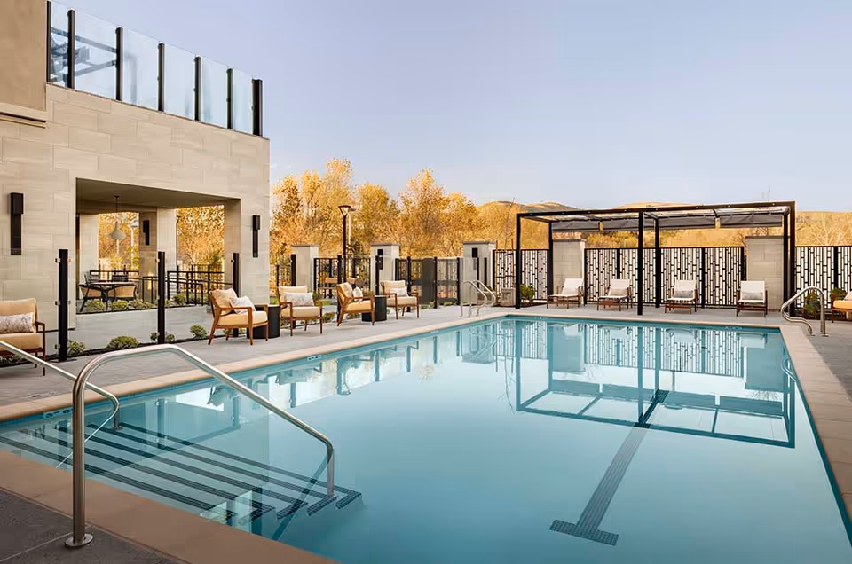 Outdoor swimming pool area with clear blue water, surrounded by lounge chairs and seating areas. There is a covered patio with additional chairs and tables, and decorative fencing in the background with trees and hills visible beyond.