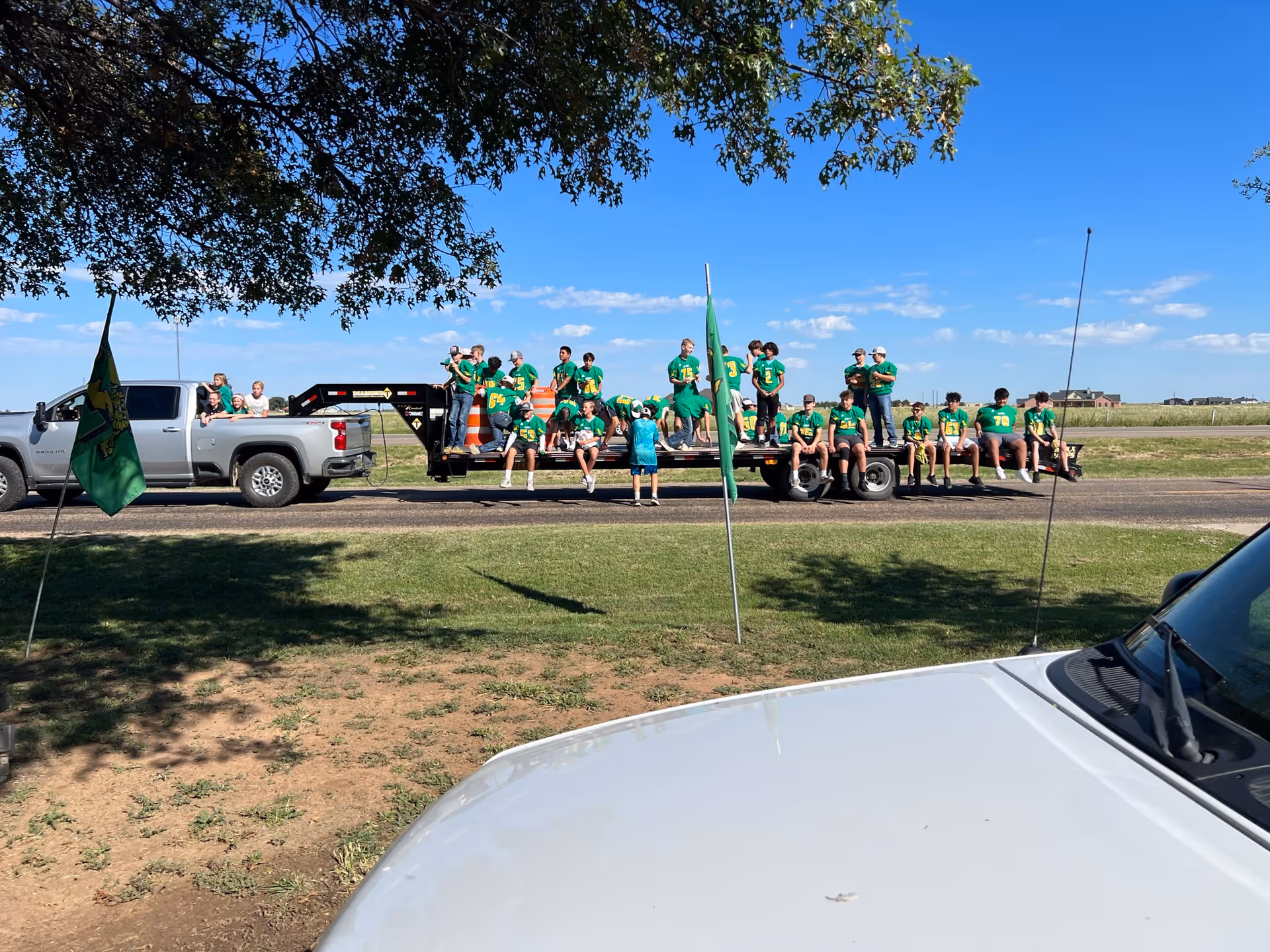 A group of people, mostly children and teenagers wearing green shirts, sitting and standing on a flatbed trailer attached to a silver pickup truck parked on a rural road. There are green flags on poles in the foreground, a white vehicle partially visible in the bottom right corner, and a large tree providing shade from above. The sky is clear and blue with a few clouds.