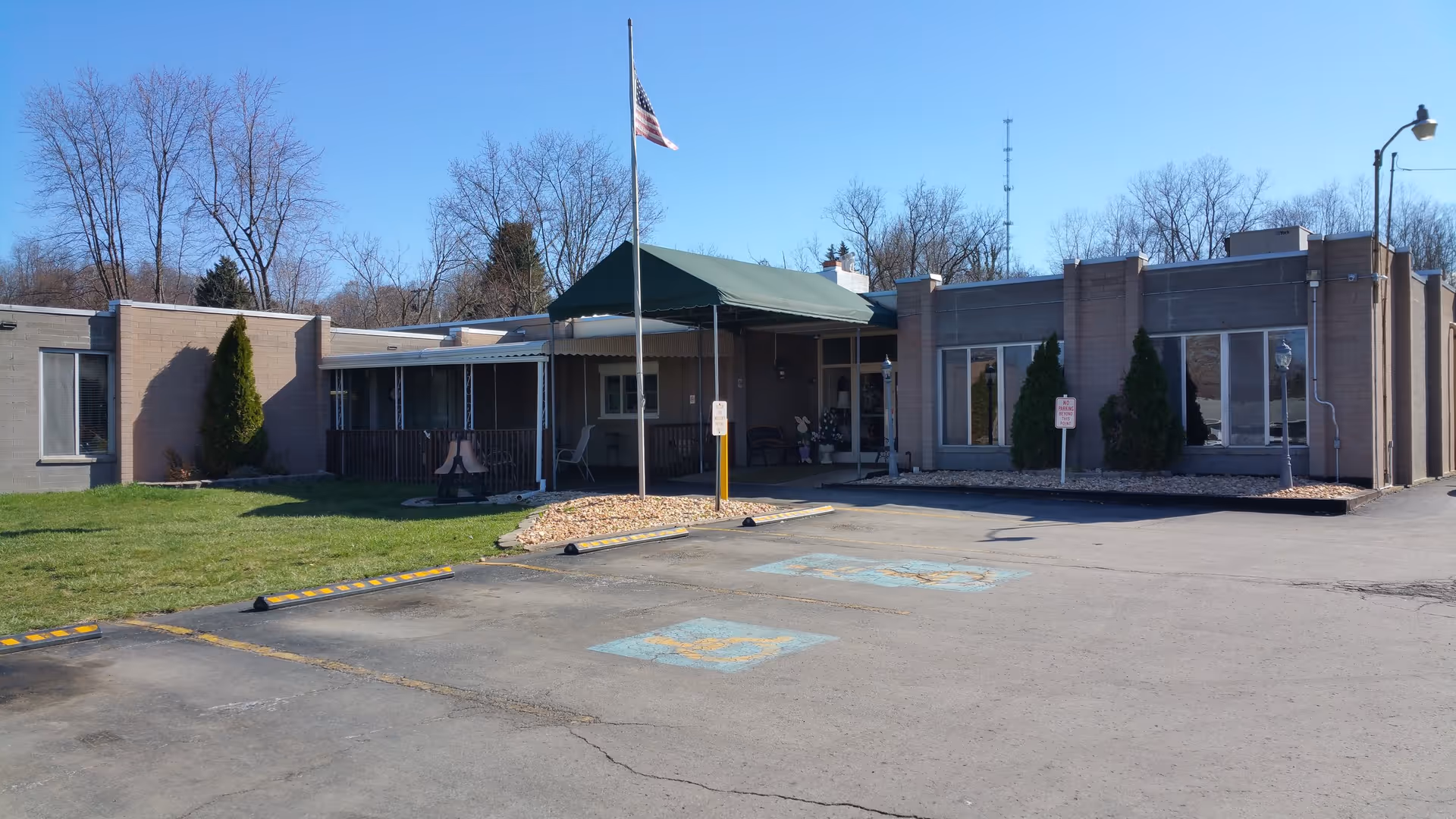 Single-story beige brick building with a covered main entrance and flagpole, a front parking area with faded handicap markings, and trees in the background.