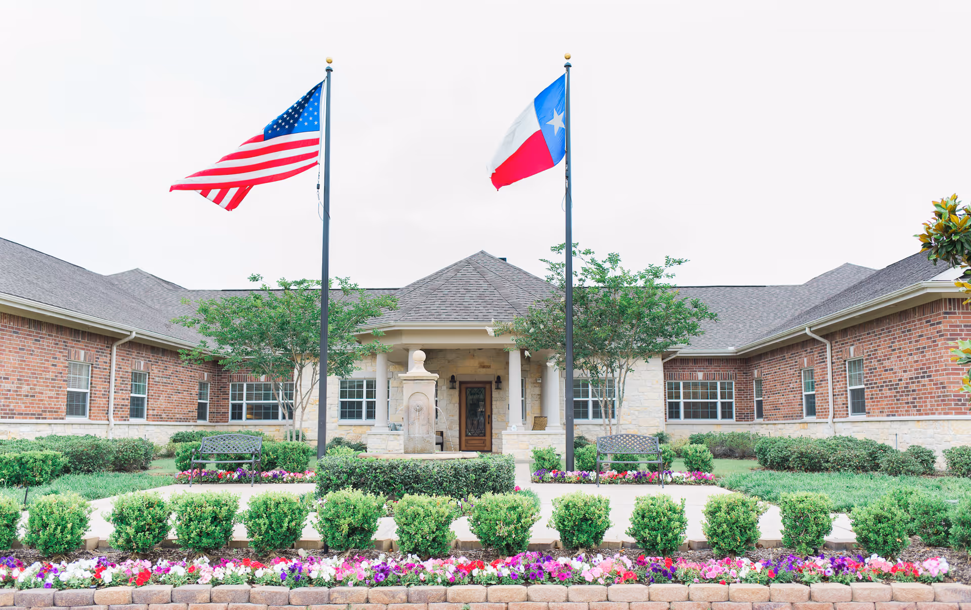Front exterior view of a single-story brick building with a central entrance, two flagpoles displaying the American and Texas flags, surrounded by well-maintained landscaping including bushes, flowers, and trees, with benches near the entrance.