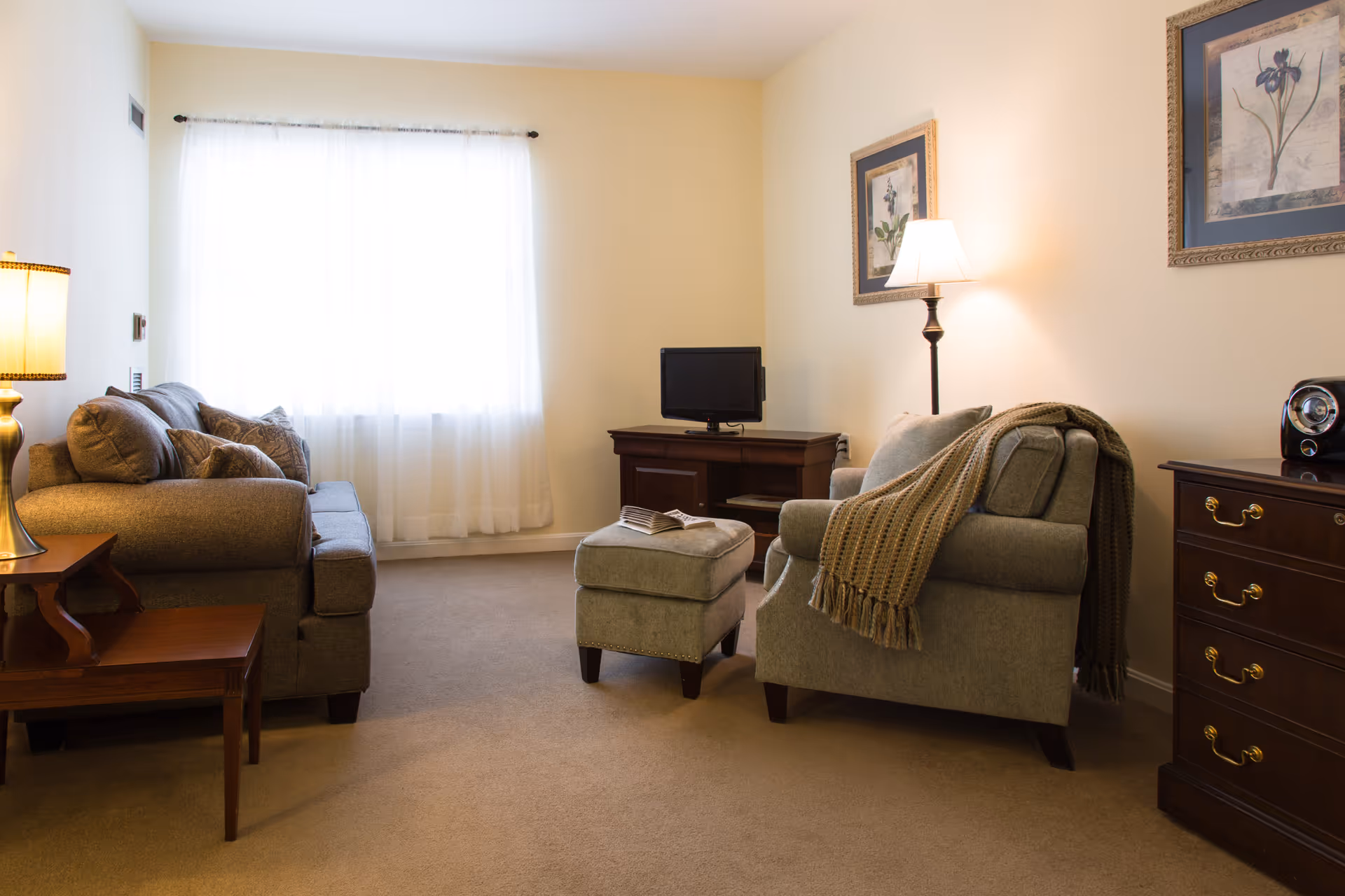 A cozy living room with beige walls and carpet, featuring a gray sofa with cushions on the left, a matching armchair with a knitted throw draped over it, and a small ottoman in the center. There is a wooden side table with a lamp on the left, a wooden TV stand with a small television in the back, and a wooden chest of drawers on the right. Two framed floral artworks hang on the walls, and a floor lamp provides additional lighting.