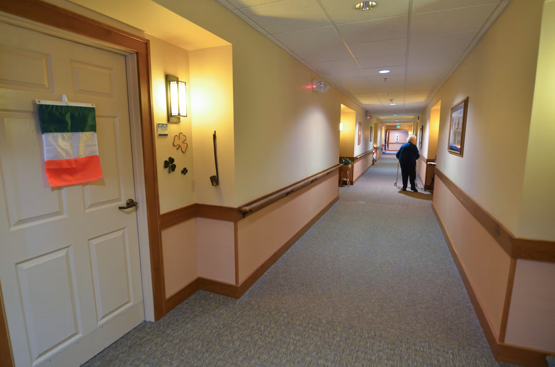 A long, well-lit hallway in a senior living facility with beige walls and carpeted floor. Handrails run along both sides of the corridor. A door on the left has an Italian flag decoration and shamrock wall decorations nearby. An elderly person using a walker is seen walking down the hallway.
