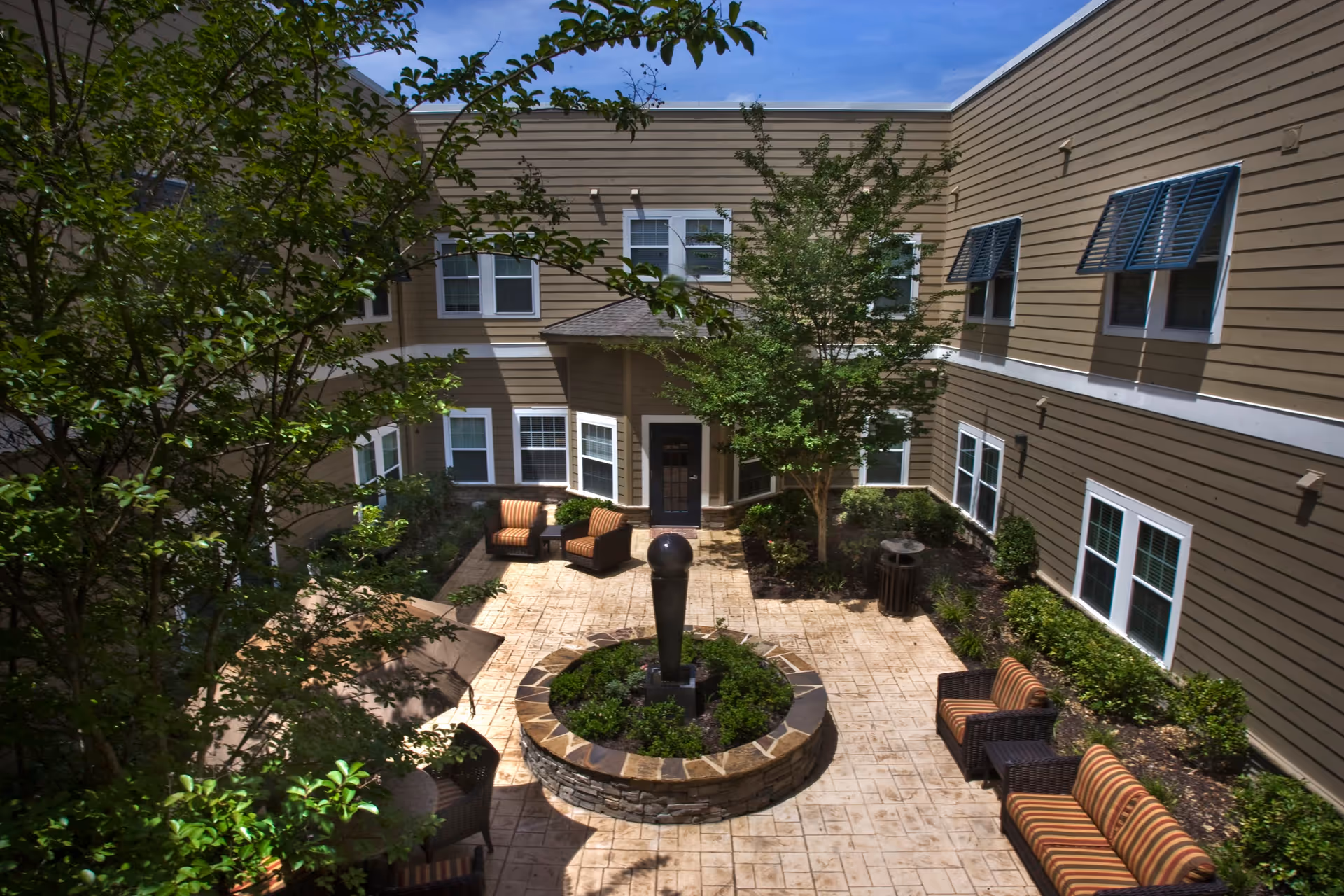 Outdoor courtyard area of a senior living facility with patio furniture including chairs and a sofa arranged around a central circular stone planter with a sculpture. The courtyard is surrounded by a two-story building with multiple windows and some trees and shrubs providing greenery.