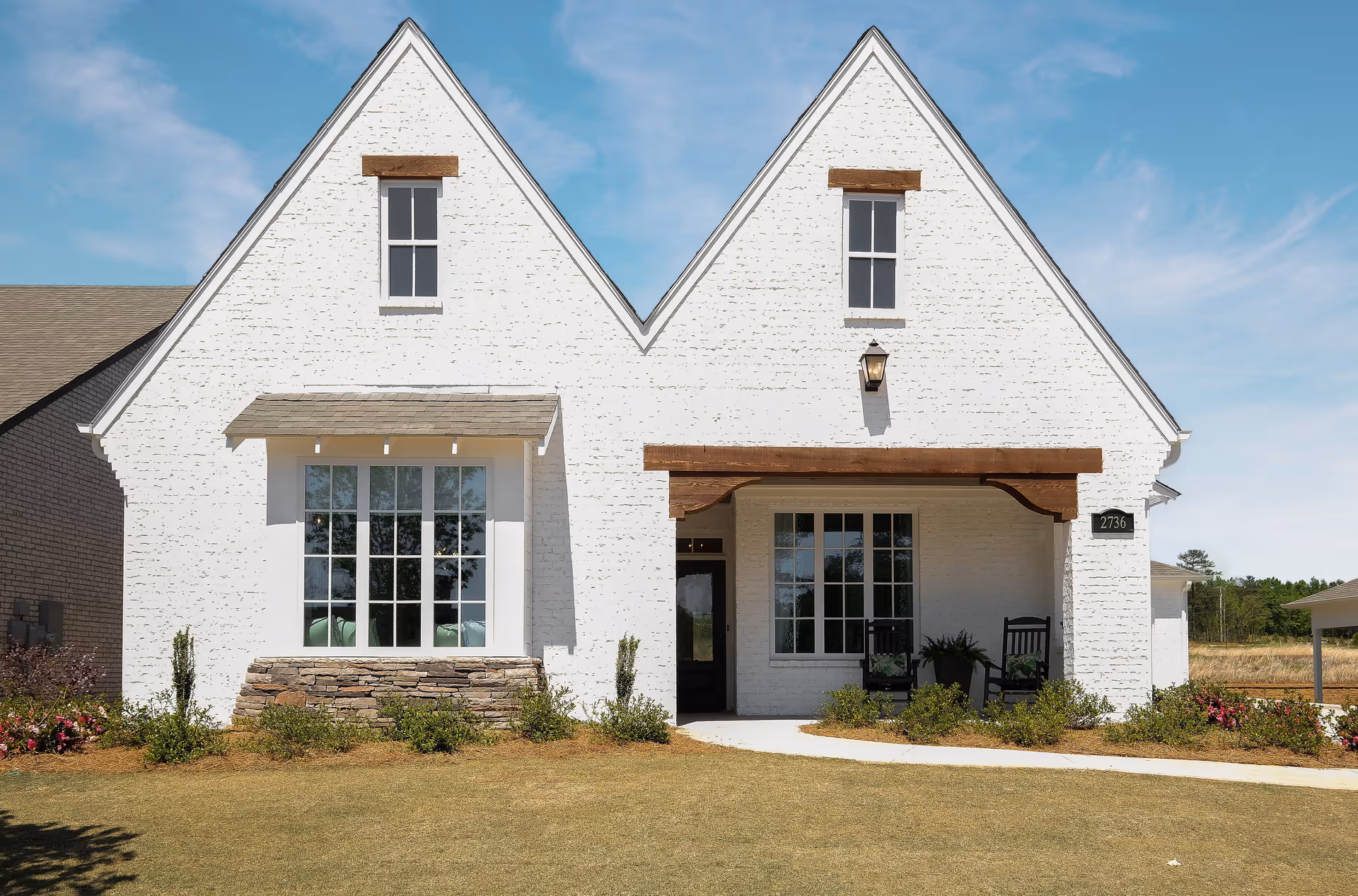 Front exterior view of a white brick building with two steep gable roofs, large windows, a covered porch with two rocking chairs, and a small garden in front under a clear blue sky.