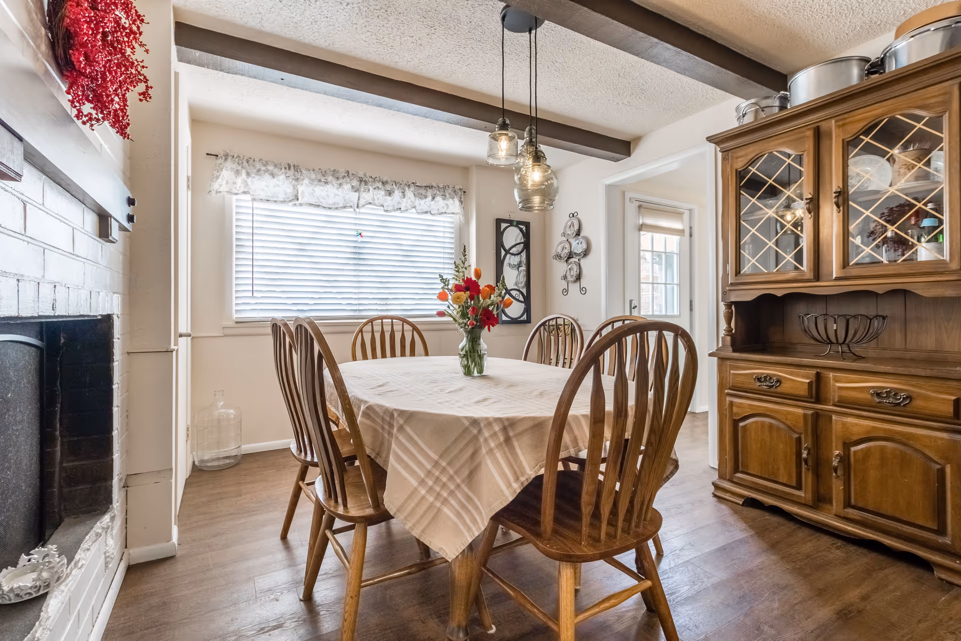 Dining room with a round table covered by a plaid tablecloth, wooden chairs, a vase of flowers, a china cabinet, and a fireplace.