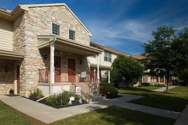 Exterior view of a residential building with stone and siding facade, featuring a small porch with stairs and red railings, surrounded by green lawns and trees under a blue sky with some clouds.