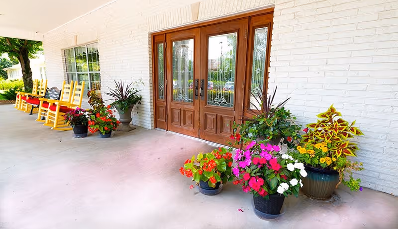 Entrance area of a building with double wooden doors featuring decorative glass panels. Several colorful potted plants are arranged near the doors. To the left, there are yellow rocking chairs and a bench placed along the wall under a covered porch.