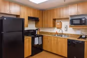 A kitchen area with wooden cabinets, a black refrigerator, black stove with oven, black dishwasher, and a black microwave. The countertop is granite with a sink and a soap dispenser mounted on the wall above it. There is a paper towel dispenser and a telephone on the counter.