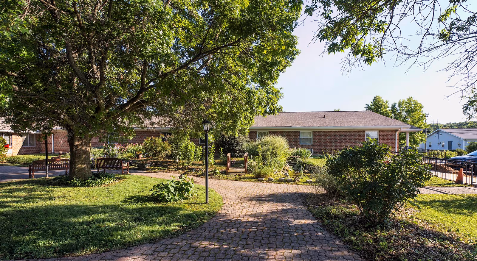 Brick single-story building with a paved courtyard path, benches, trees, and landscaped greenery in front.