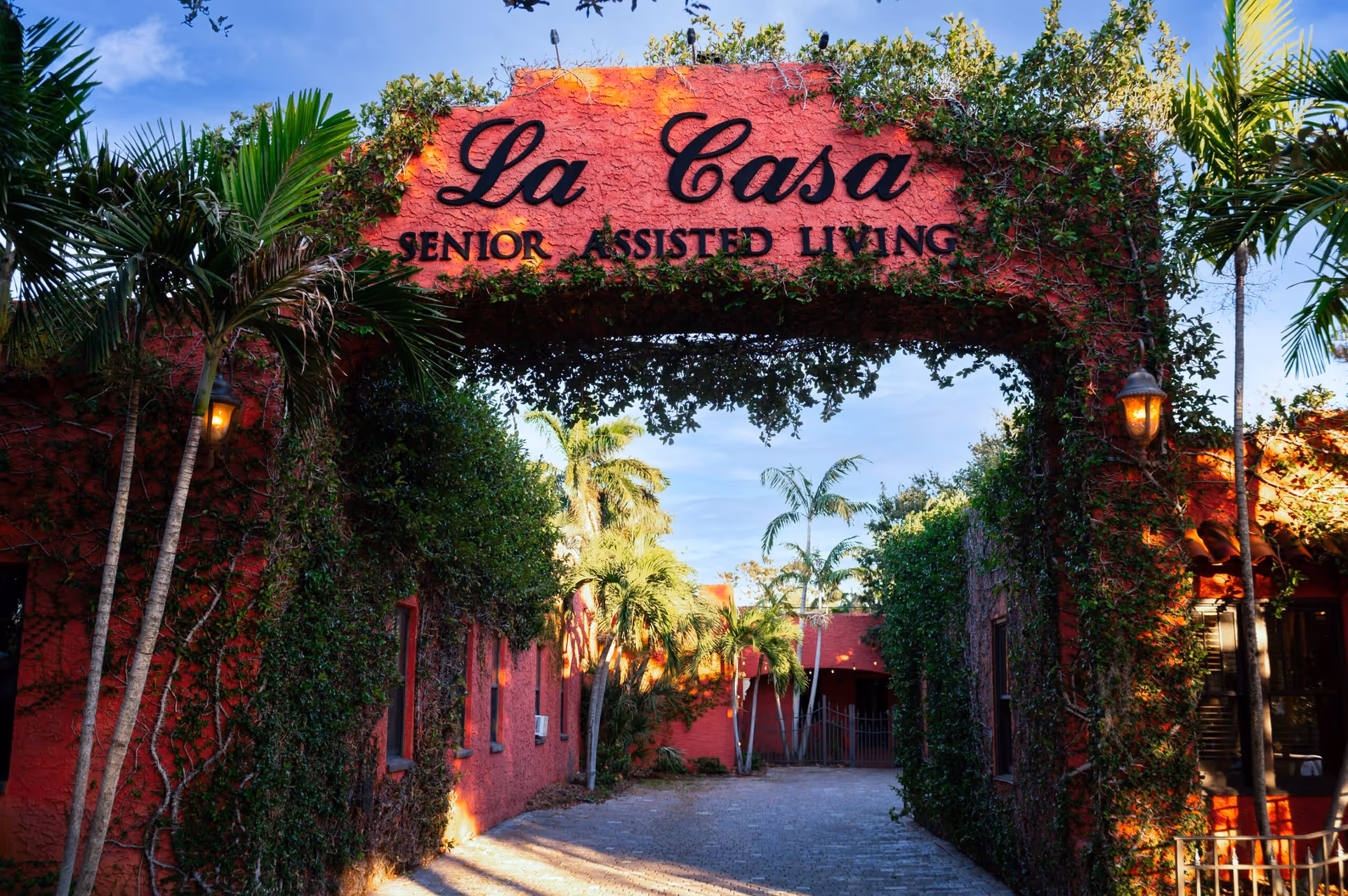 Entrance archway covered with green ivy and palm trees on either side, with the text 'La Casa Senior Assisted Living' displayed on the arch. The building and surrounding walls are painted red, and there is a paved pathway leading inside under a clear blue sky.