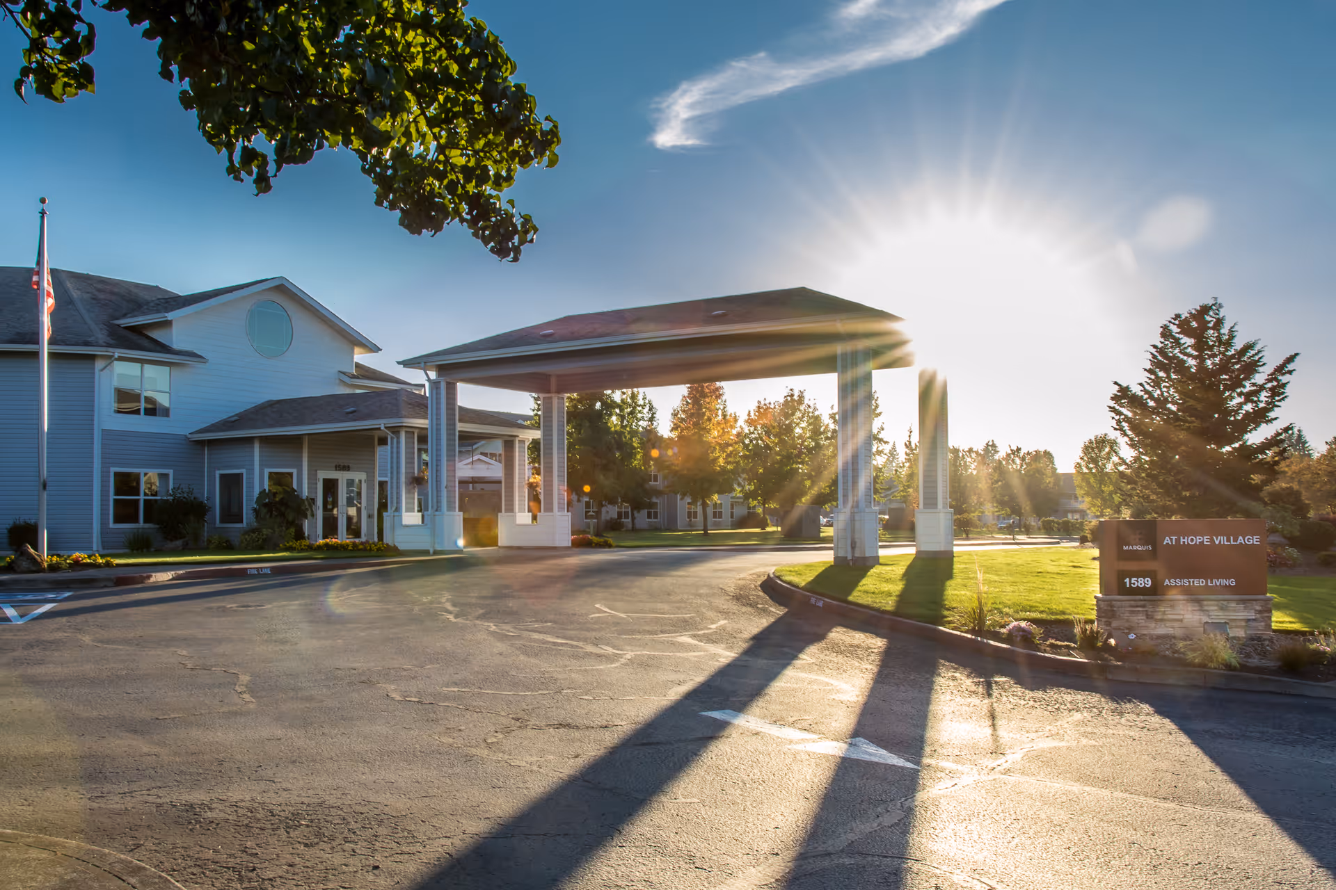 Assisted living facility front entrance with a covered drop-off canopy, sun flare, landscaping, and a sign reading 'At Hope Village'.