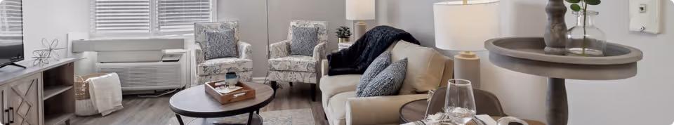 A cozy living room featuring a beige sofa with patterned cushions, two matching armchairs, a round wooden coffee table with a tray, a TV on a wooden stand, and a window with blinds. The room has wooden flooring and neutral-colored walls with modern decor.
