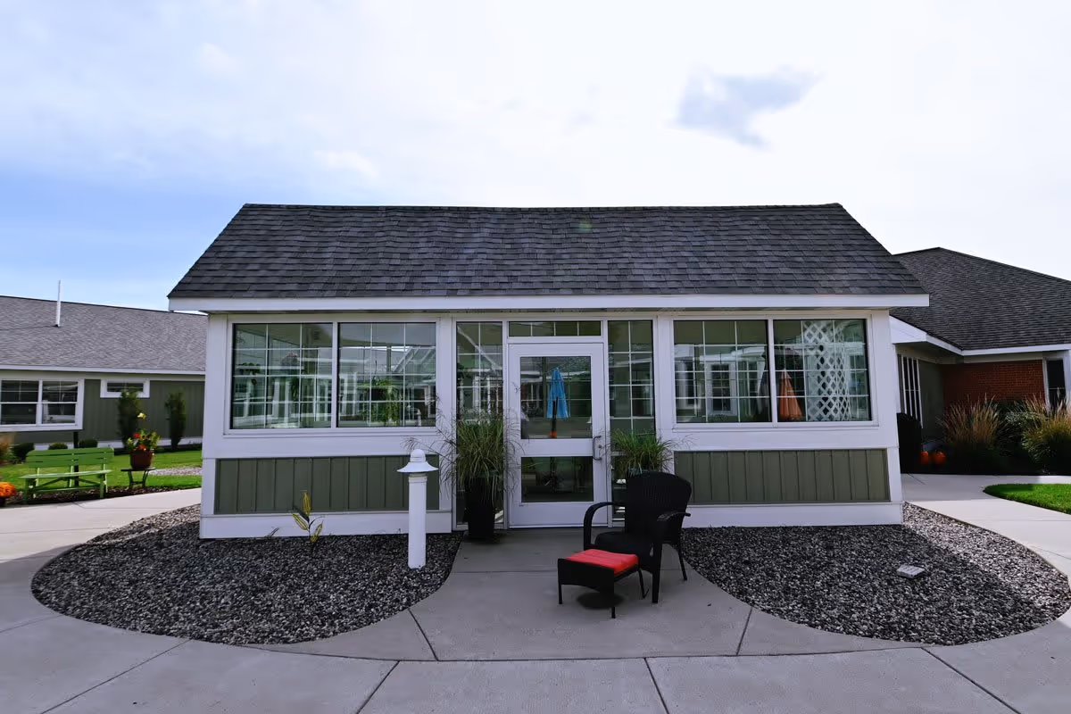 Exterior view of a small building with large windows and a door in the center. There is a black chair with a red cushion and a matching footrest on the concrete pathway in front of the building. The building has a shingled roof and green and white siding. Surrounding the pathway are landscaped areas with rocks and some plants. Other buildings and green benches are visible in the background under a partly cloudy sky.