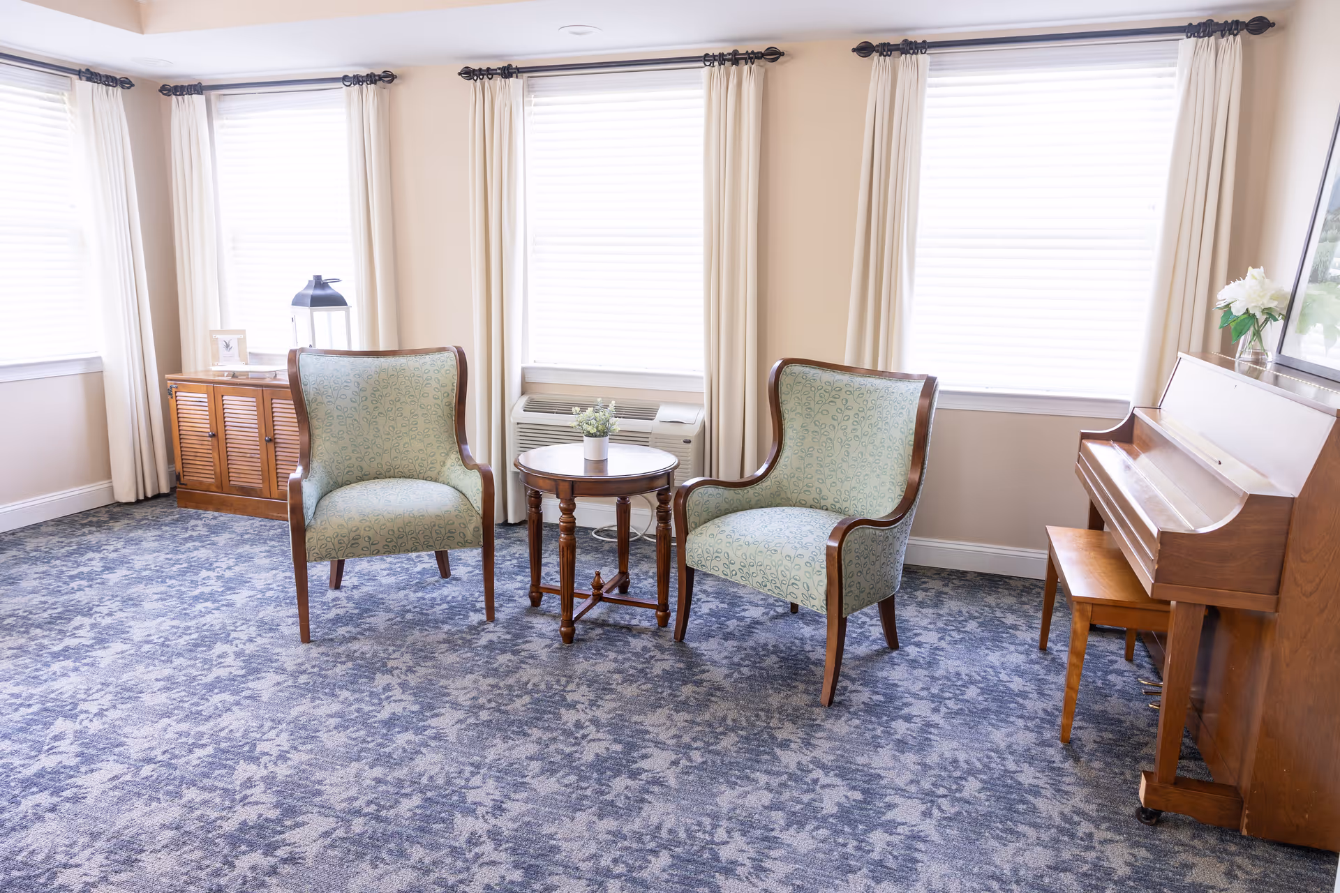 A bright room with three large windows covered by white blinds and cream curtains. Two patterned armchairs with wooden frames are positioned around a small round wooden table with a small potted plant on it. To the right, there is a wooden upright piano with a matching bench and a vase of white flowers on top. On the left side, there is a wooden cabinet with a lantern and a framed picture on it. The floor is covered with a blue patterned carpet.