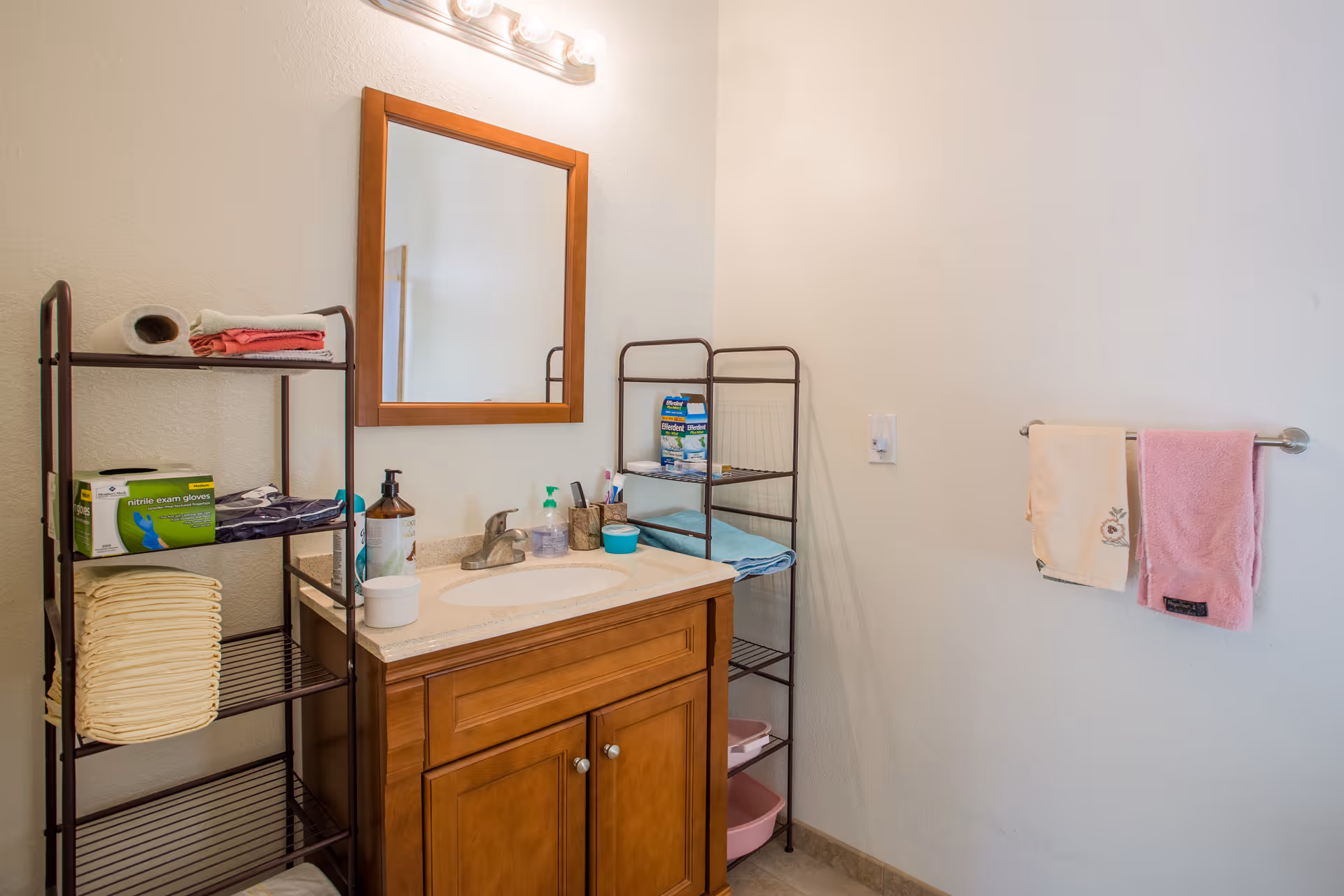 A bathroom with a wooden vanity and sink, a rectangular mirror above it, and two metal shelving units holding towels, gloves, and other supplies. Two towels hang on a wall-mounted towel rack to the right.