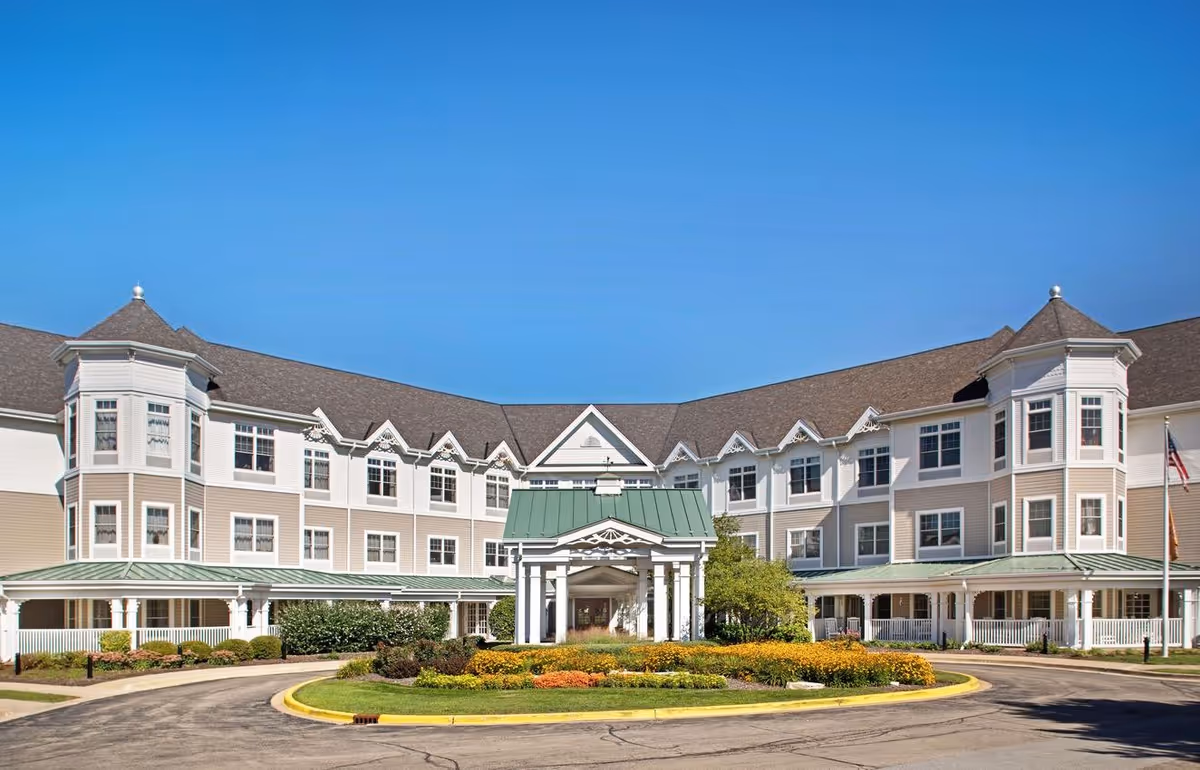 Front view of a large three-story senior living building with a covered entrance, landscaped circular driveway, and clear blue sky.